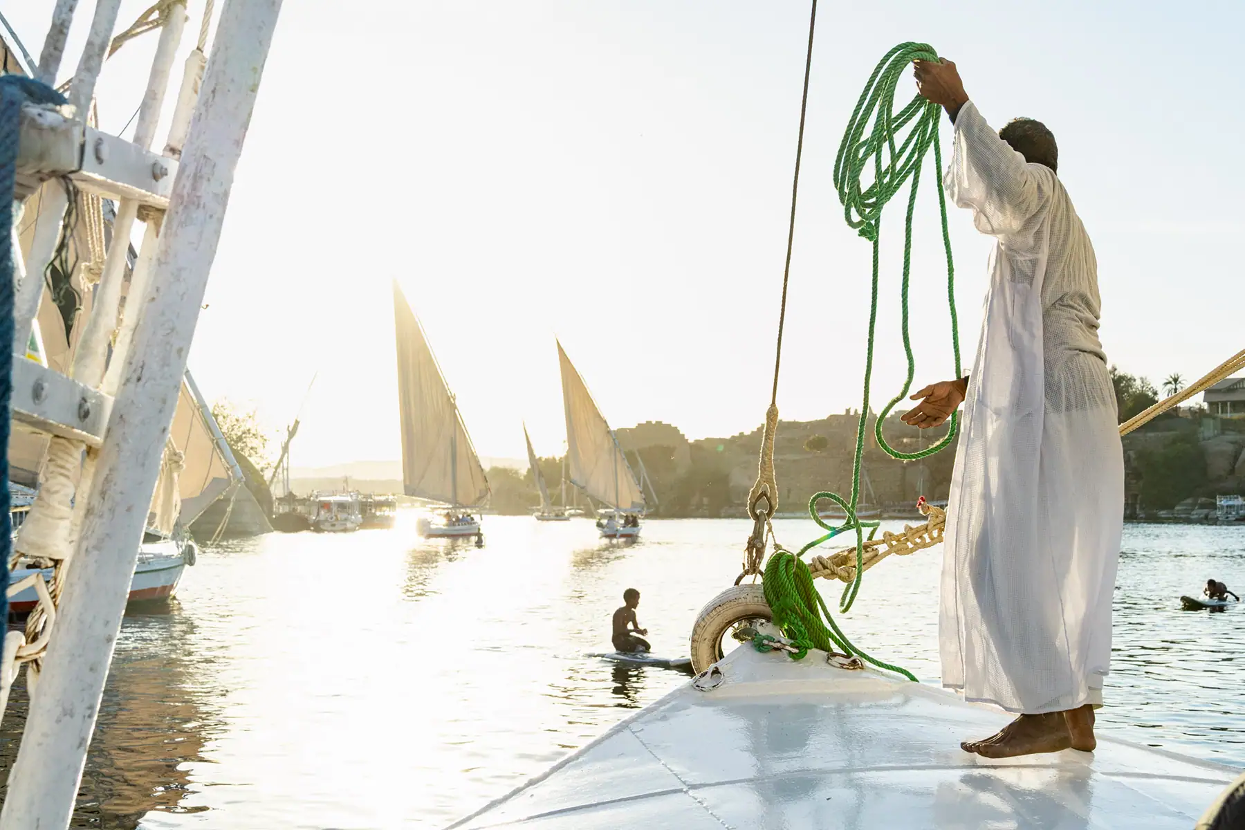 A local sailor handles a green rope aboard a felucca at sunset on the Nile River, with traditional sailboats and swimmers in the background for a unique cultural moment to photograph.