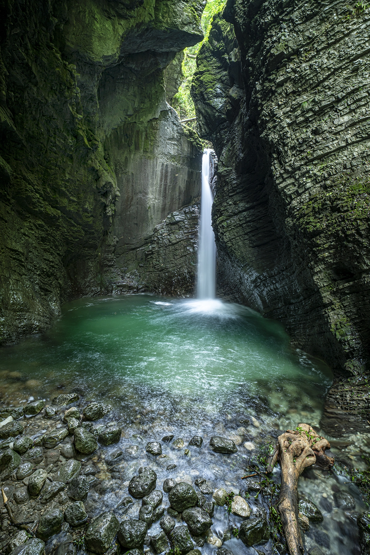 A secluded waterfall plunging into a crystal-clear emerald pool, surrounded by towering moss-covered cliffs and rocky terrain.