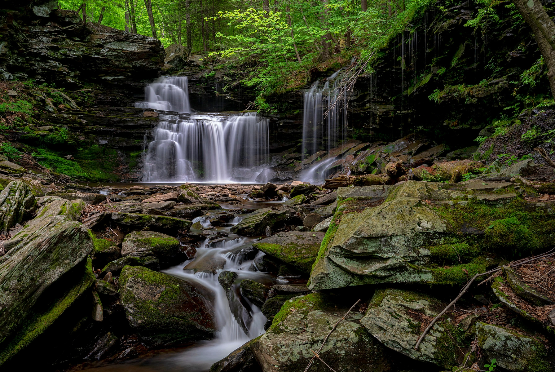 A cascading waterfall flowing over layered rocks, surrounded by lush green foliage and moss-covered stones.