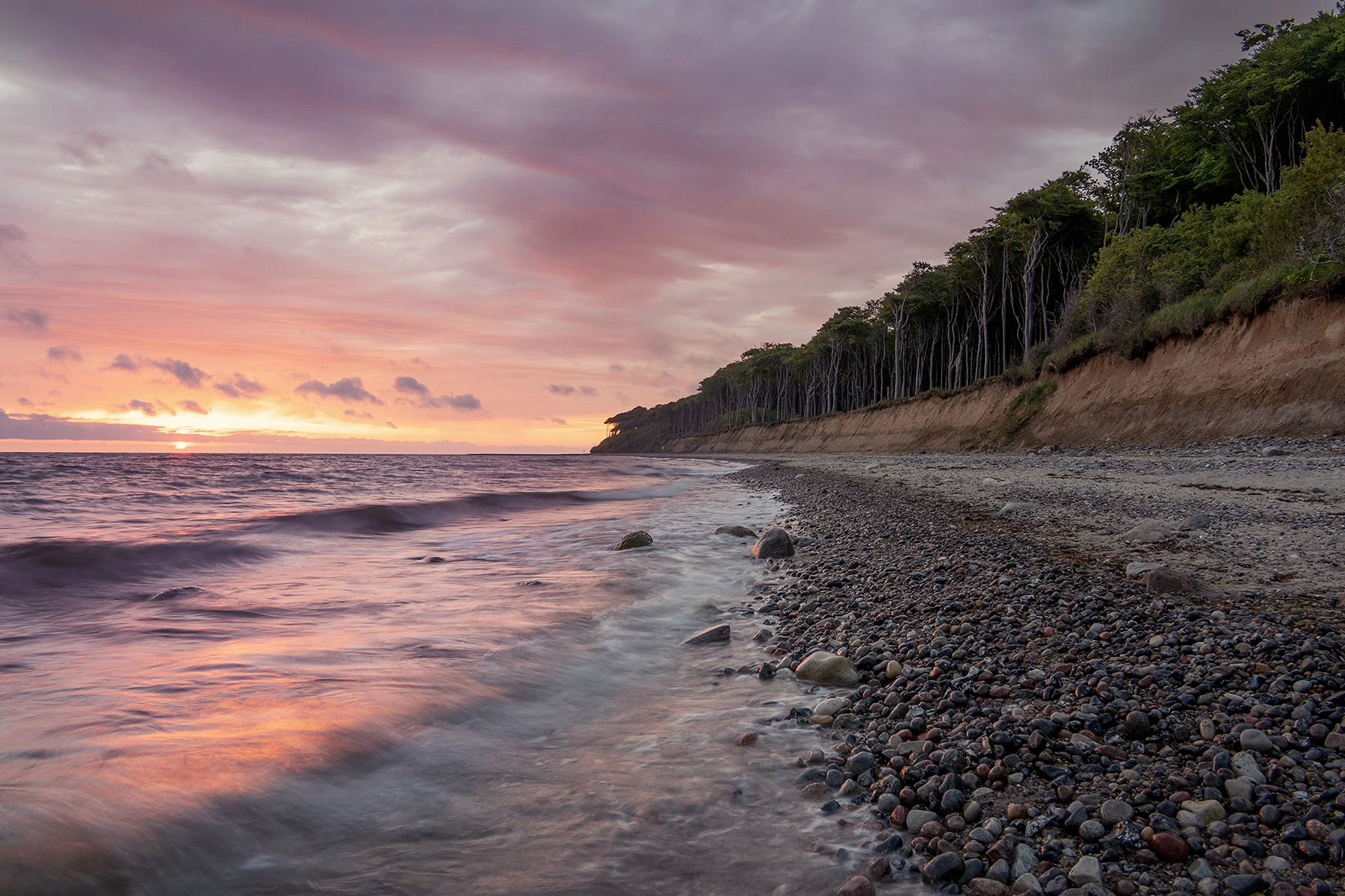 A serene beach at sunset with gentle waves lapping against a pebble-covered shoreline, framed by dramatic cliffs and a vibrant, colorful sky.
