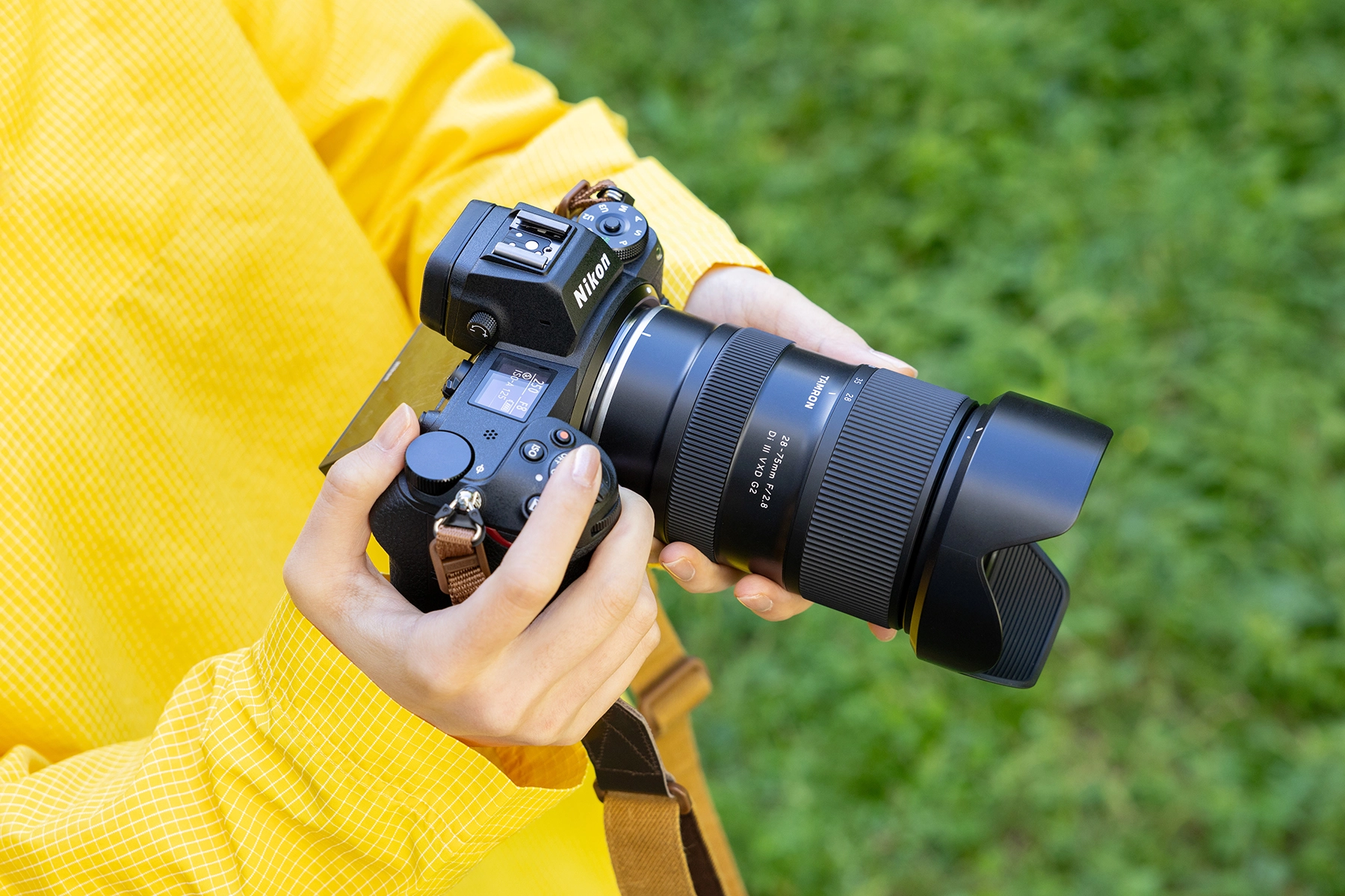 Photographer holding a Nikon Z series mirrorless camera with a compact and lightweight TAMRON 28-75mm F/2.8 lens, ready for shooting in a green outdoor setting.
