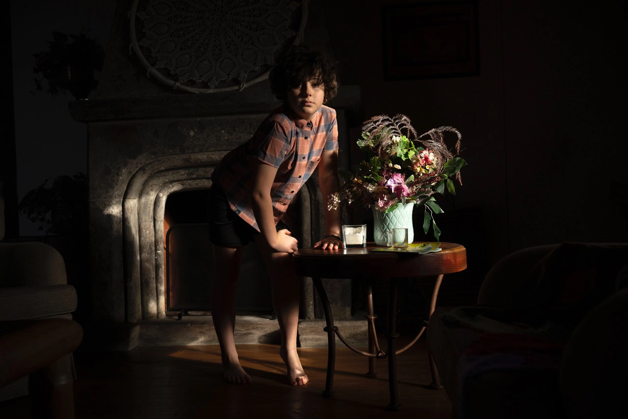 A moody indoor portrait of a child lit by early morning light, standing near a table with a floral arrangement&mdash;an example of photographing portraits in early morning light using natural window illumination.