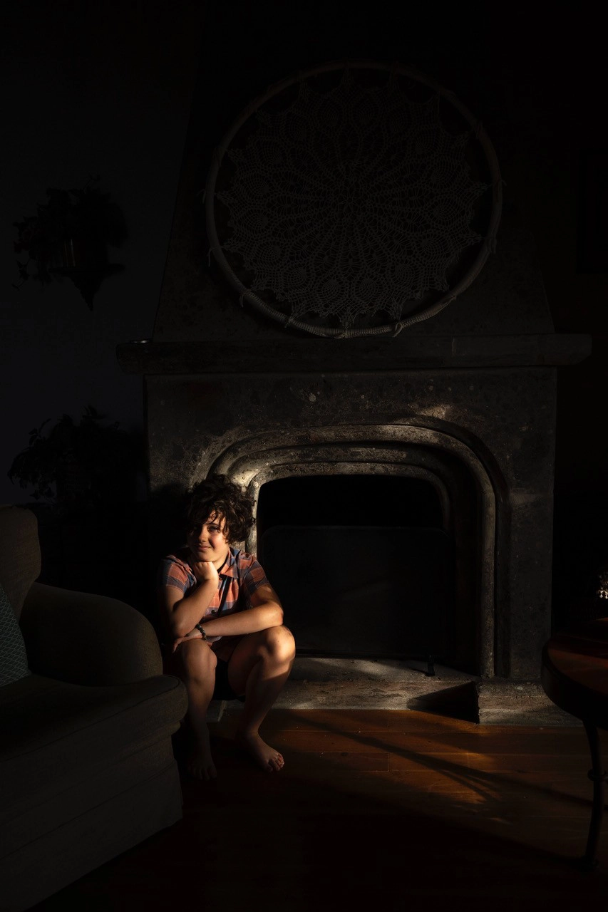 A young girl seated by a fireplace with a soft beam of early morning sunlight illuminating the face&mdash;an atmospheric example of photographing portraits in early morning light with dramatic shadows.