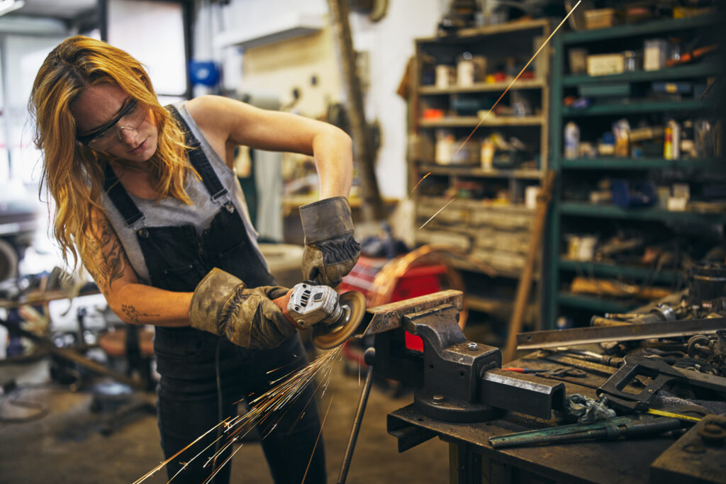 A female metalworker in gloves and goggles grinds steel in a workshop as sparks fly&mdash;demonstrating how to get a cinematic look using dynamic motion, warm tones, and rich detail in an industrial setting.