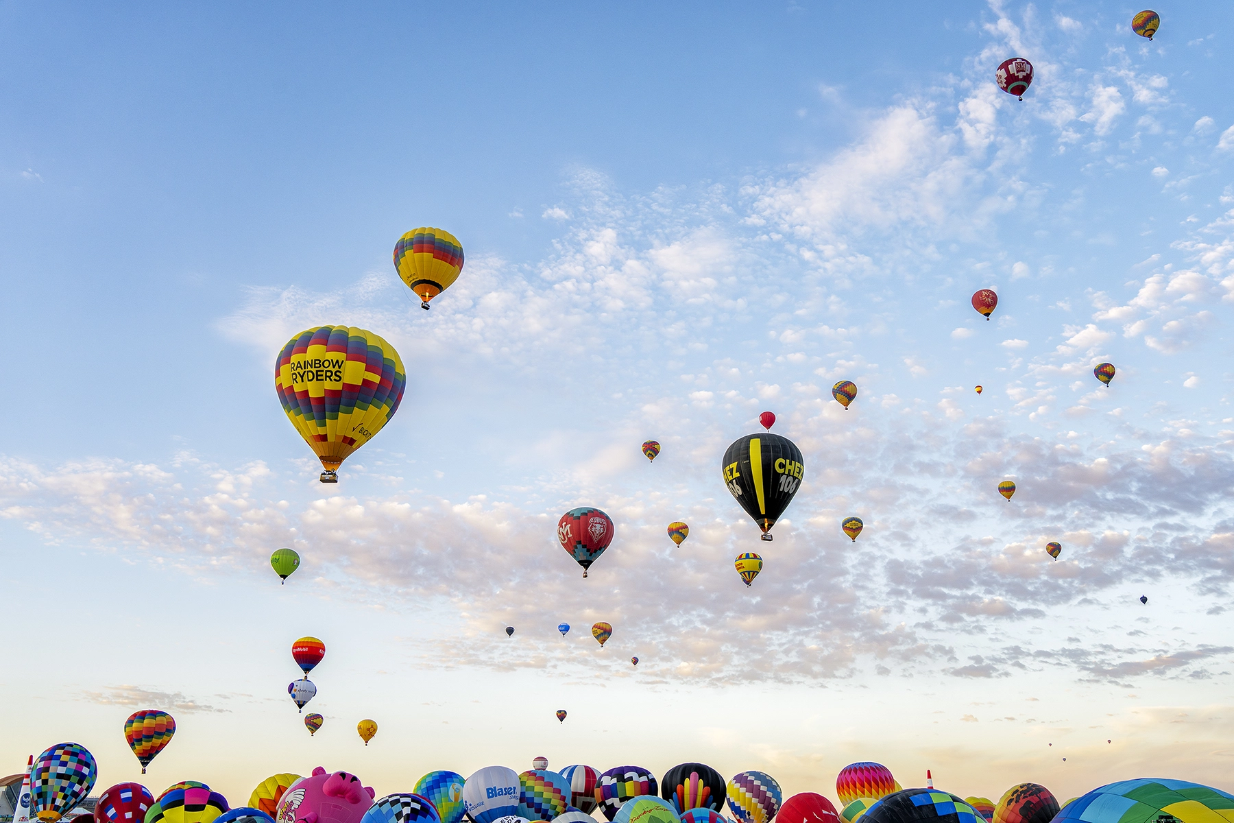 Globos aerostáticos de varios colores y diseños ascendiendo en un cielo parcialmente nublado durante un festival de globos, con el globo "Rainbow Ryders" en un lugar destacado.