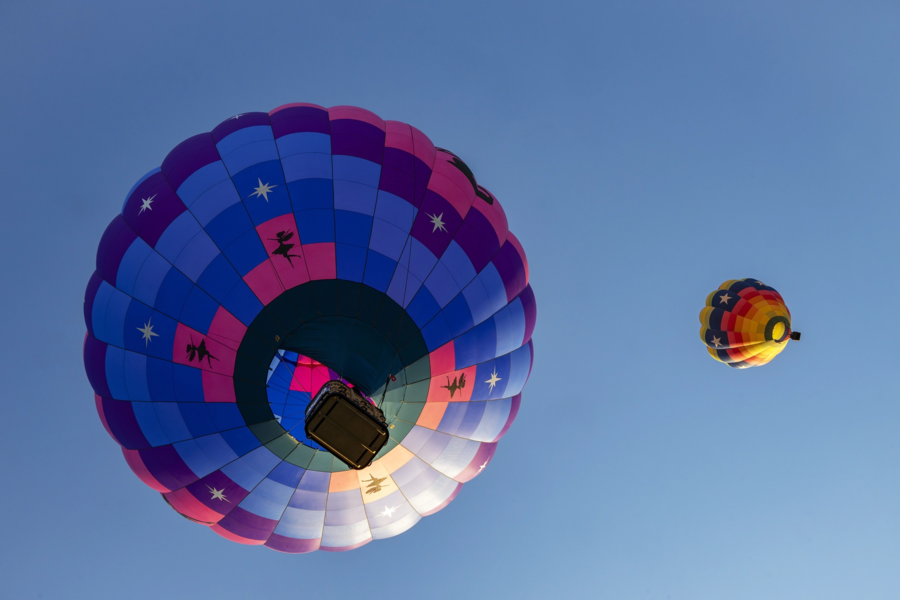 Dos globos aerostáticos de colores vistos desde abajo contra un cielo azul despejado, uno con dibujos de estrellas y siluetas y el otro con un motivo de arco iris.
