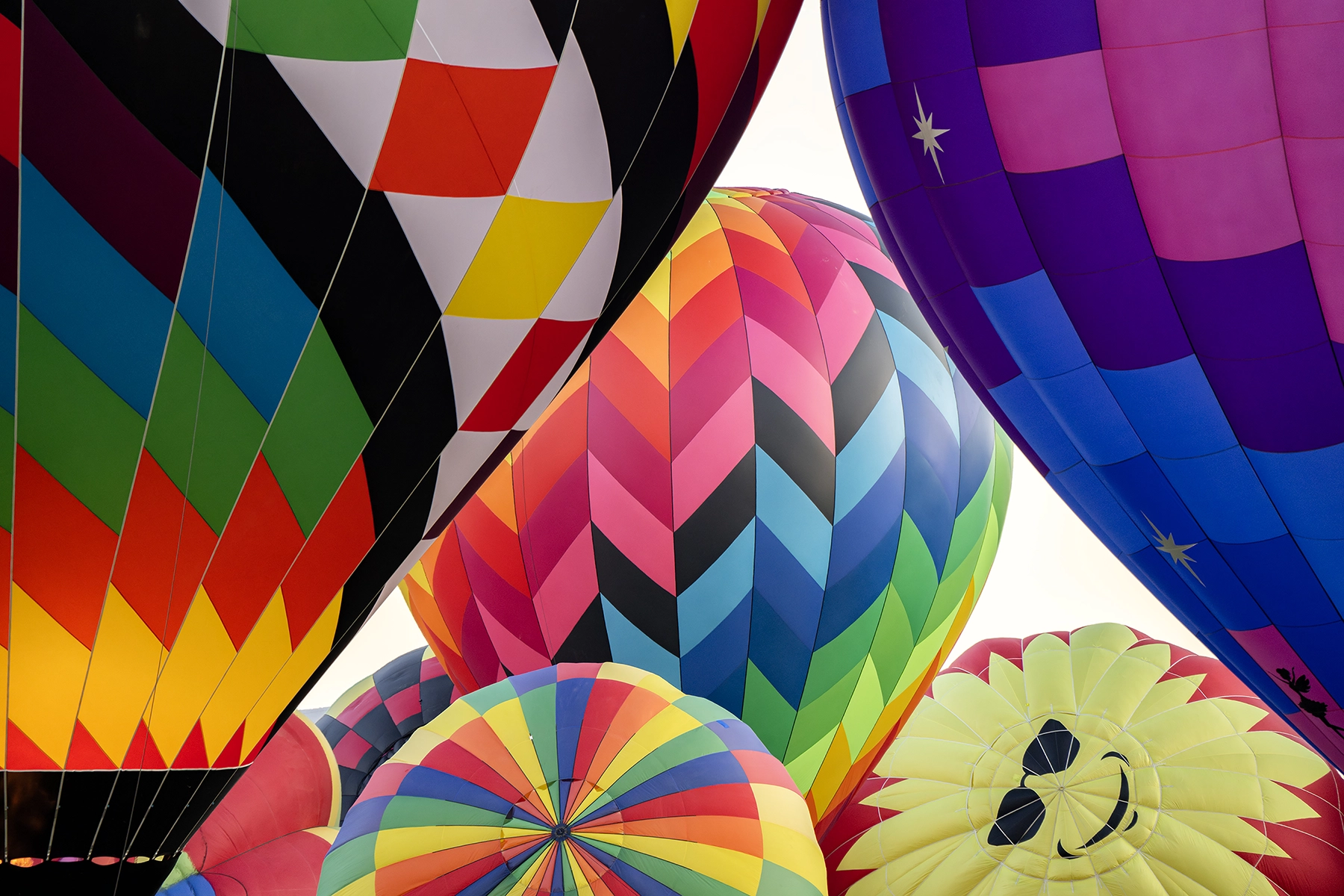 Primer plano de globos aerostáticos de colores con diversos motivos, como rayas, galones y un sol sonriente, agrupados en un festival de globos.