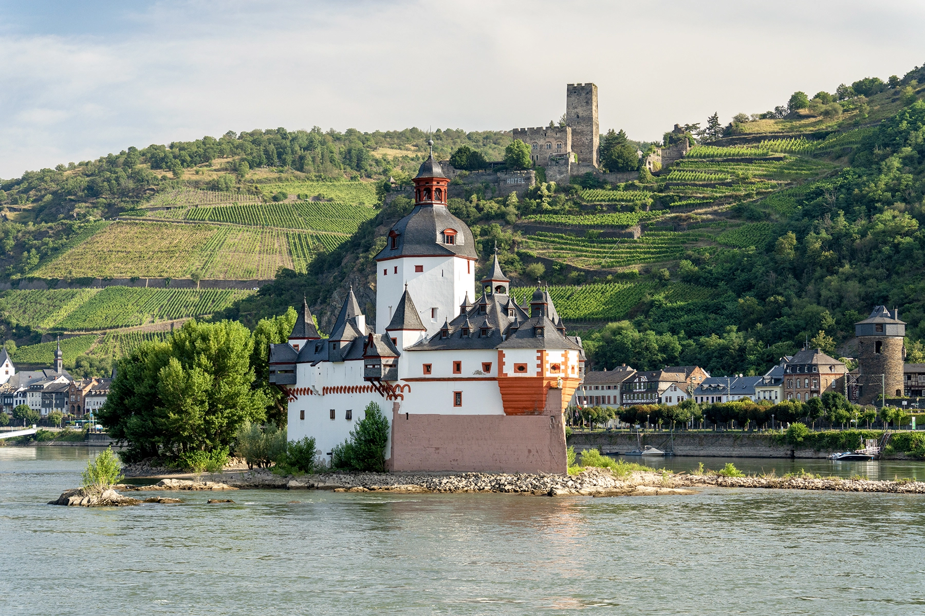 Pfalzgrafenstein Castle on a small island in the Rhine River, with vineyards and a hilltop castle in the background. This image epitomizes Rhine River cruise travel photography.