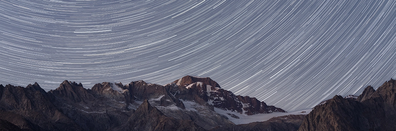 Long-exposure star trails arc above rugged alpine peaks and glacial terrain, captured with a 90mm macro lens for detailed night sky photography.