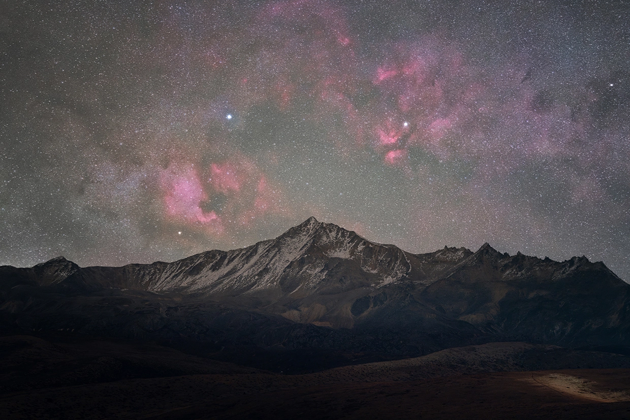 The North America and Pelican Nebulae glow vividly above a jagged mountain ridge, revealed in striking clarity through astrophotography with a macro lens.
