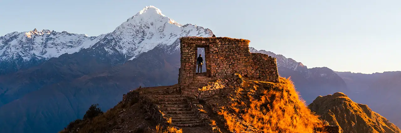 A photographer framed in a stone ruin overlooking a dramatic mountain range at golden hour—captured with a camera lens for mountain photography to emphasize detail, elevation, and warm light.