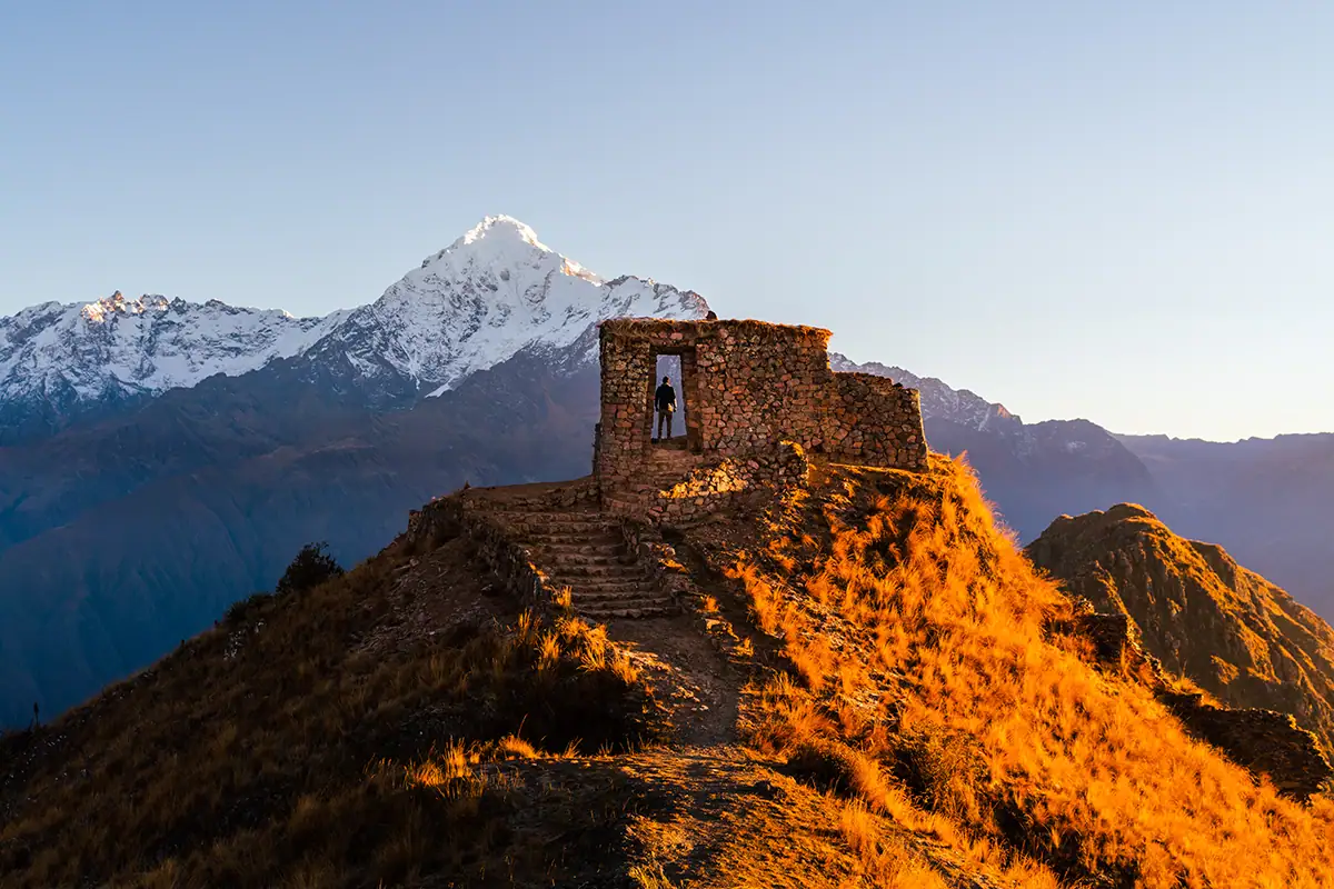 A lone traveler stands inside a stone ruin at sunrise, framed by golden alpine light and a snow-capped mountain backdrop&mdash;captured with a camera lens for mountain photography to emphasize texture, scale, and elevation.