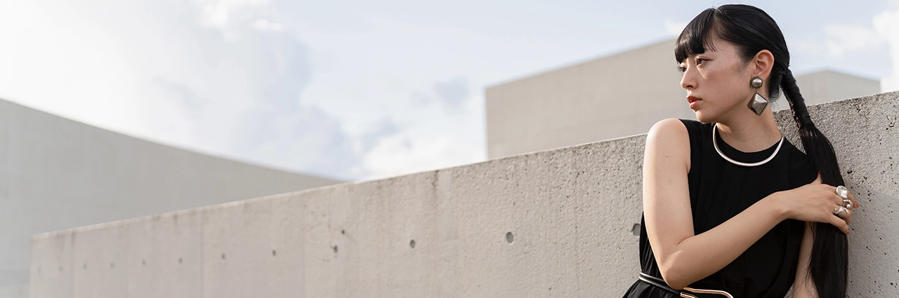 A woman with long dark hair styled in braids, wearing a black sleeveless dress and silver jewelry, leans against a textured concrete wall with a modern building and a cloudy sky in the background.