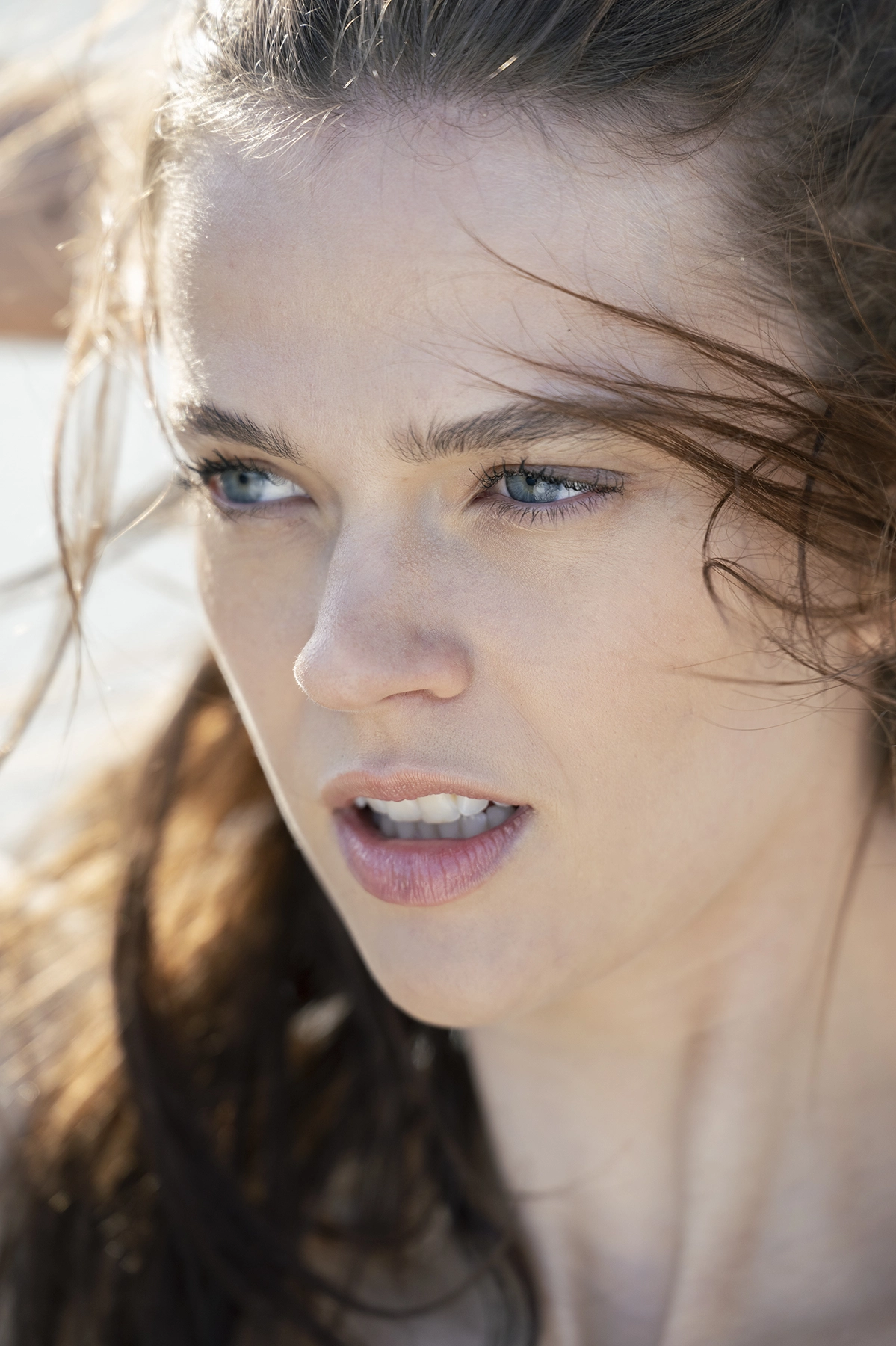 A close-up portrait of a woman with blue eyes and windswept brown hair, gazing into the distance with a determined expression under natural sunlight.
