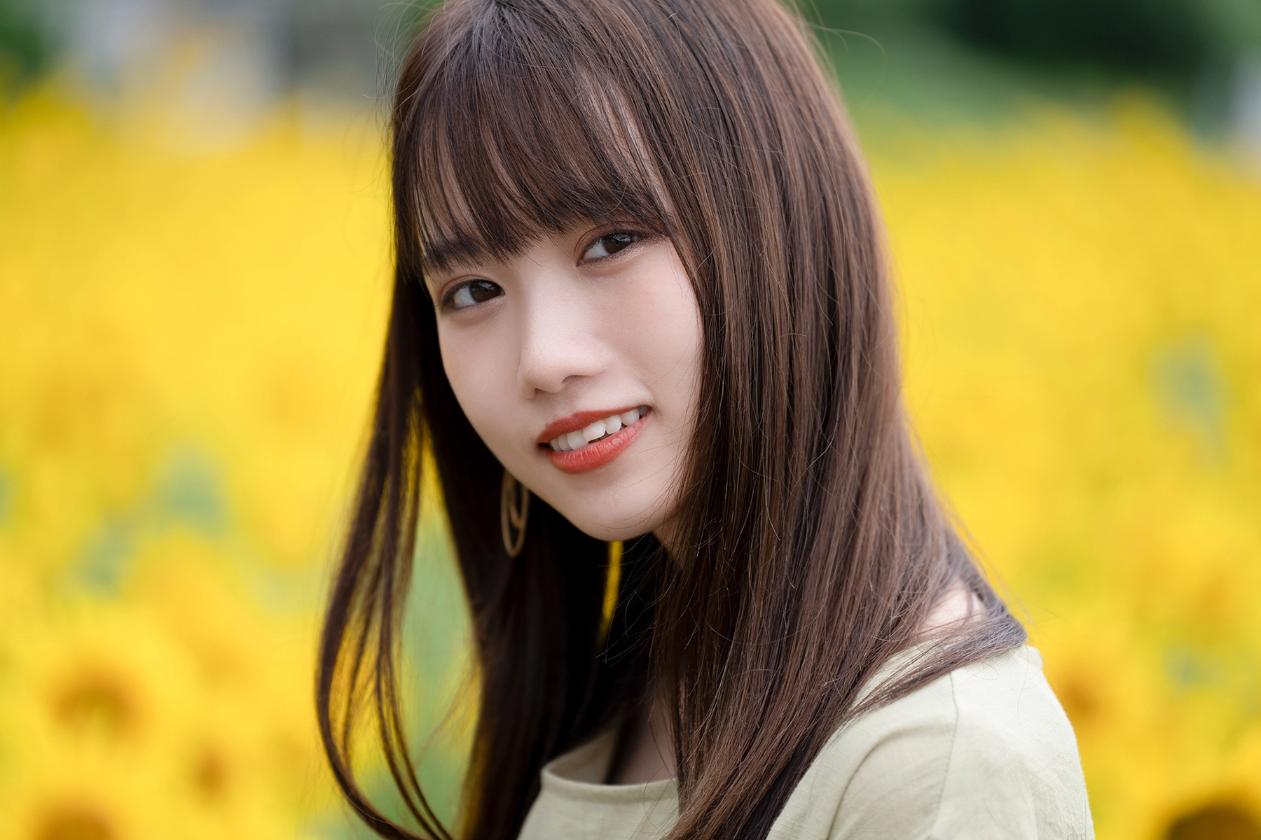 A young woman with long, straight brown hair and a gentle smile poses in front of a vibrant field of yellow flowers, with a soft, blurred background.