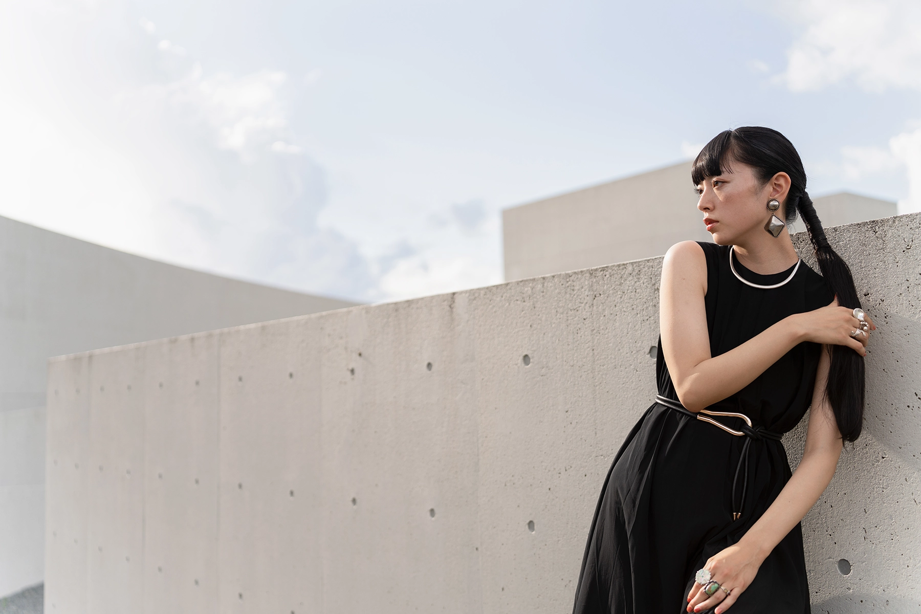 A woman with long dark hair styled in braids, wearing a black sleeveless dress with a metallic belt and statement jewelry, leans against a textured concrete wall with a modern, minimalist building and a cloudy sky in the background.