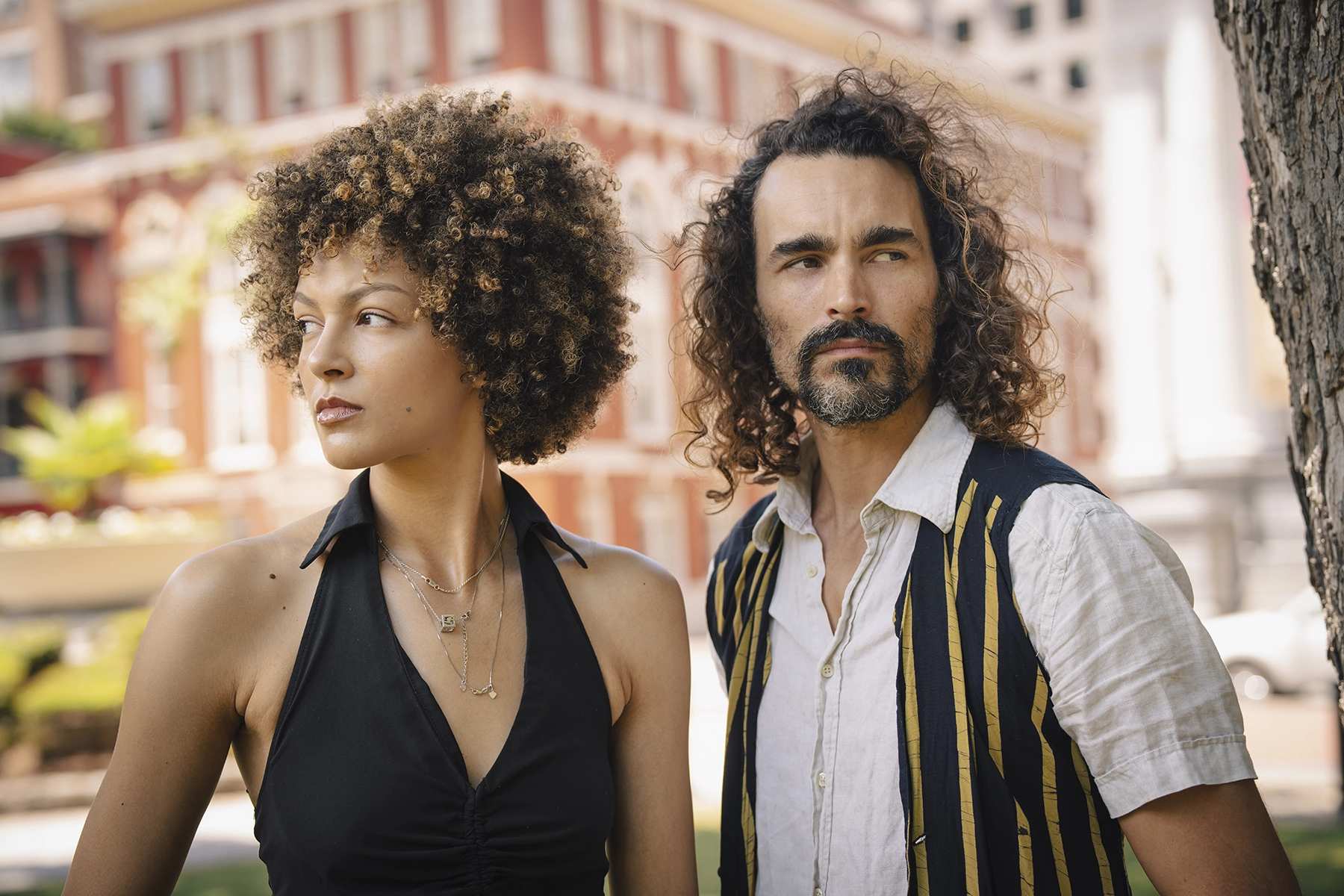 A woman with curly hair wearing a black halter top and layered necklaces stands beside a man with long curly hair and a beard, wearing a striped vest over a white shirt, both gazing off to the side with a historic red-brick building in the background. Photographed with a wide aperture makes the subjects pop off the background.