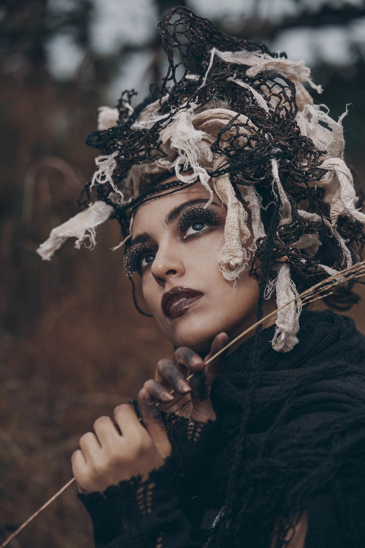 A woman with dramatic makeup and dark lips gazes upward, wearing an intricate headpiece made of tattered fabric and netting, with her hands gently holding dried reeds against a blurred natural background.