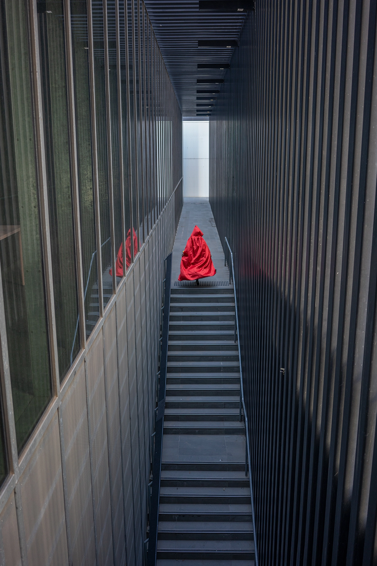 Persona con una llamativa capa roja subiendo por una escalera urbana minimalista enmarcada por líneas arquitectónicas oscuras: un momento artístico que pone de relieve cómo el mejor objetivo para fotografía de viajes capta el contraste, la simetría y el ambiente en los paisajes urbanos.