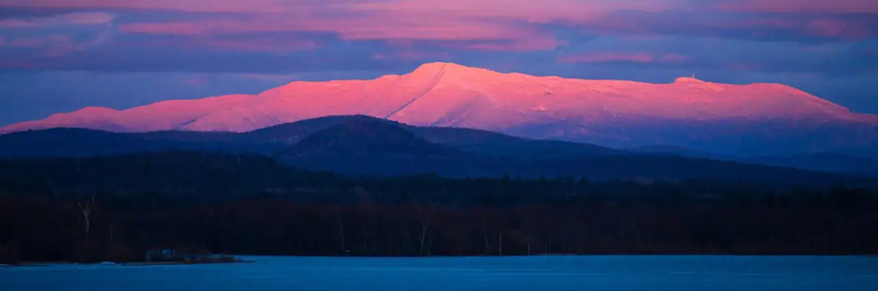 A breathtaking winter landscape featuring a snow-covered mountain bathed in soft pink and purple hues from the setting sun, with a frozen lake and dark forested hills in the foreground.
