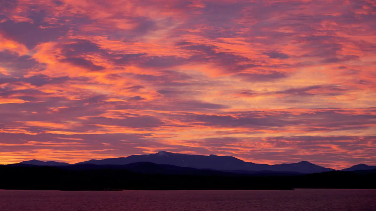 A stunning sunset over a mountain range, with vibrant shades of pink, orange, and purple filling the sky and reflecting softly on the water below.