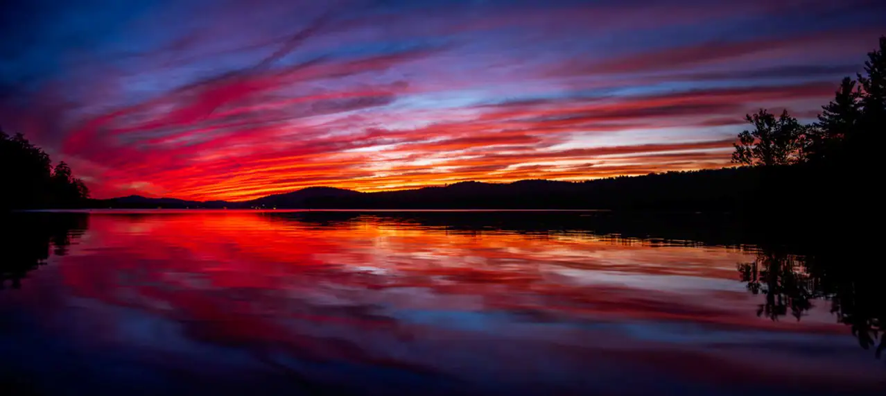 A stunning sunset over a calm lake, with vibrant red, orange, and purple hues streaking across the sky and reflecting beautifully on the water&rsquo;s surface.