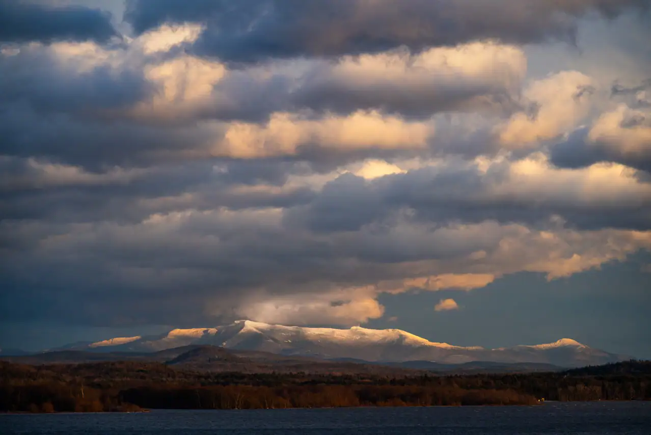 A dramatic landscape of snow-capped mountains illuminated by golden sunlight, partially shrouded in heavy clouds, with a dark lake and forest in the foreground.