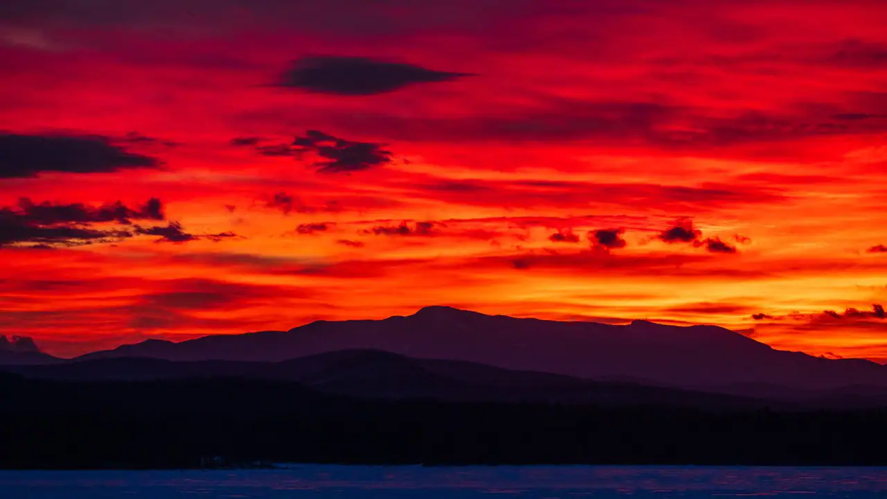 A fiery sunset ignites the sky in deep reds and oranges over a silhouetted mountain range, with dramatic clouds adding texture to the vibrant scene