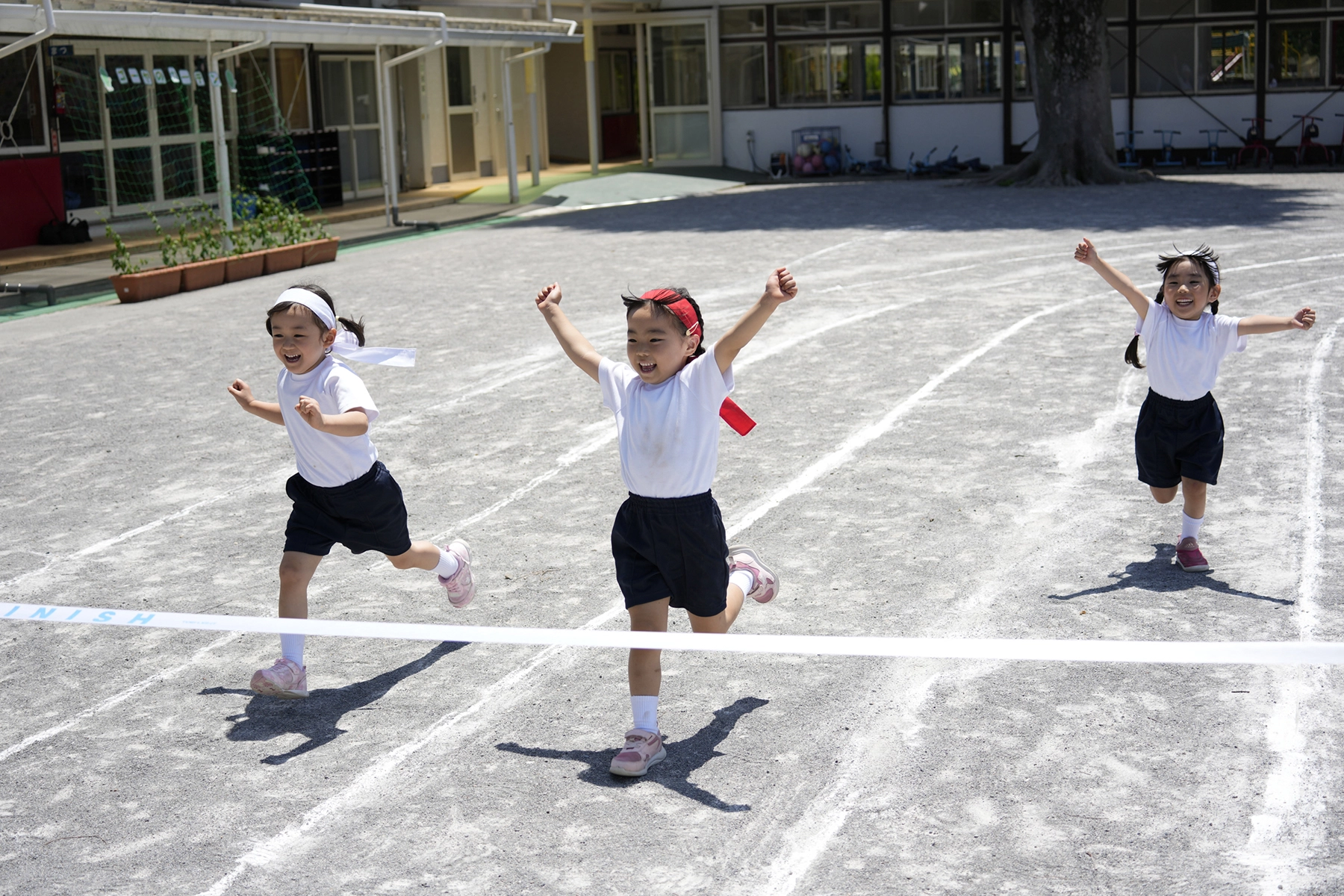 An action shot featuring three young children joyfully racing toward the finish line on a school sports day, arms raised in excitement under bright sunlight on a gravel track.