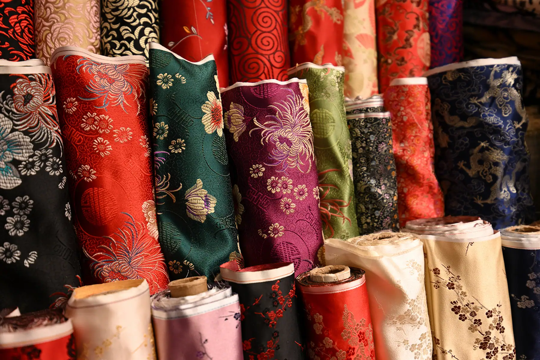Gros plan sur des rouleaux de tissu aux motifs complexes, dans une variété de couleurs et de textures vibrantes, disposés à la verticale sous une lumière douce et naturelle.