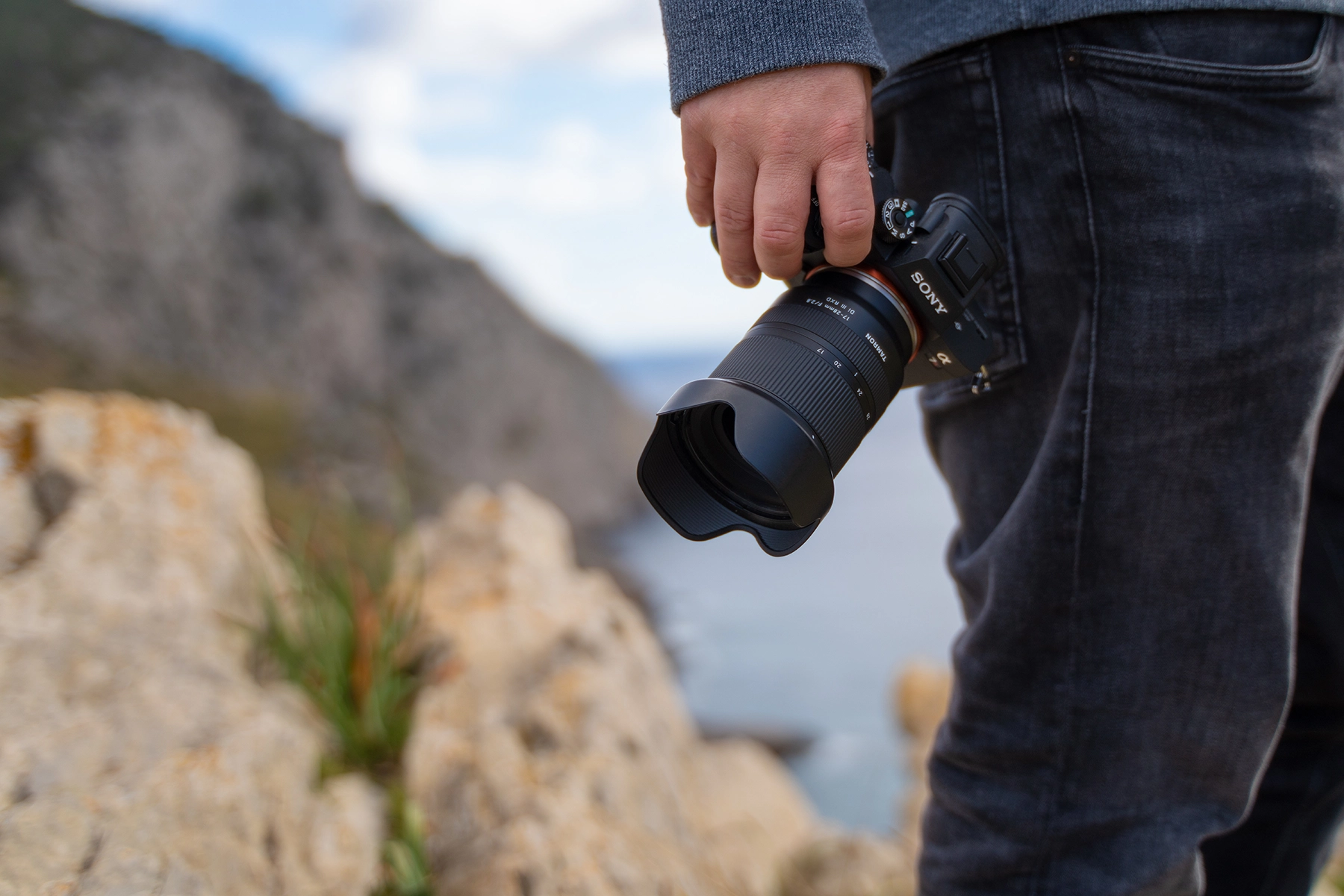 Close-up of a person holding a Sony mirrorless camera with a Tamron lens, standing on rocky terrain with a scenic background.