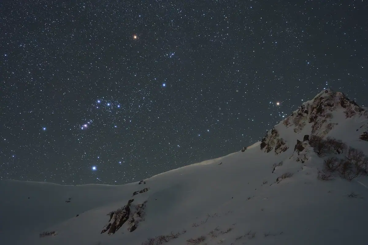 Céu noturno repleto de estrelas sobre um cume de montanha nevado com a constelação de Órion visível, capturado durante uma sessão de astrofotografia de inverno.