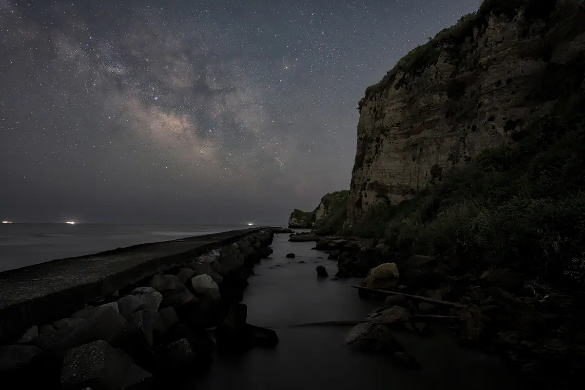 Galáxia da Via Láctea sobre um penhasco costeiro e quebra-mar, capturada durante uma paisagem noturna de longa exposição mostrando estrelas e a linha costeira em silhueta.