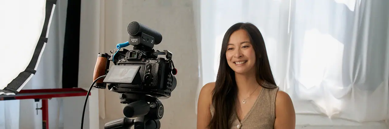 Woman sitting in front of a camera setup with an external Sennheiser microphone and Tamron lens, recording video in a softly lit studio.