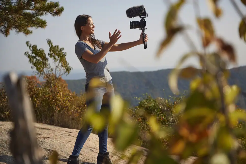 Content creator vlogging on a mountaintop using an external microphone with a mirrorless camera setup for outdoor video production.
