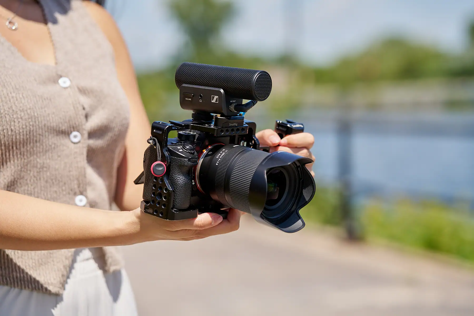 Person holding a mirrorless camera rig equipped with a Tamron lens and an external Sennheiser microphone, filming outdoors near a waterfront.