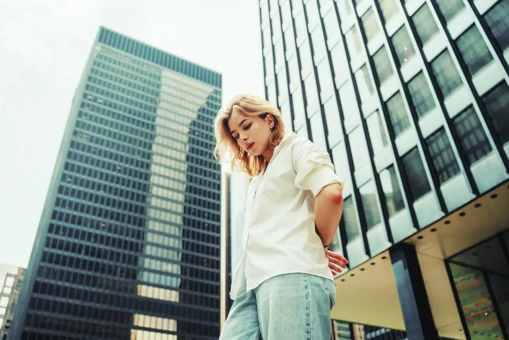 Uma mulher de camisa branca e calça jeans posando em frente a prédios altos na cidade.