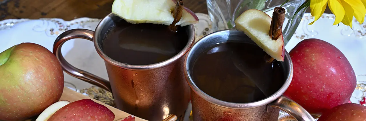 Duas canecas de cobre de cidra de maçã decoradas com fatias de maçã e paus de canela em uma mesa rústica, com estilo para fotografia de alimentos e bebidas de outono.