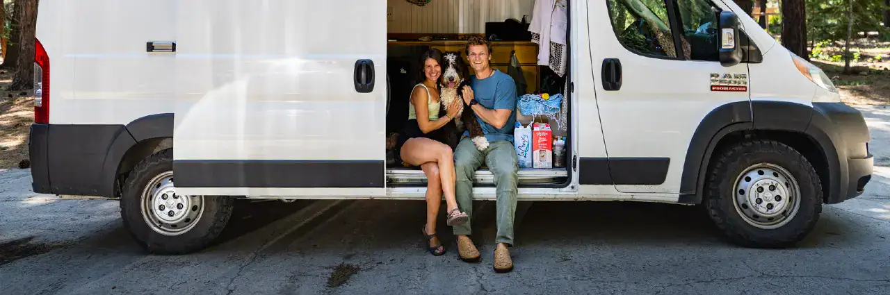 Pareja con perro sentada en la puerta de la furgoneta durante un viaje por carretera, sonriendo para el artículo de consejos fotográficos sobre viajes por carretera.
