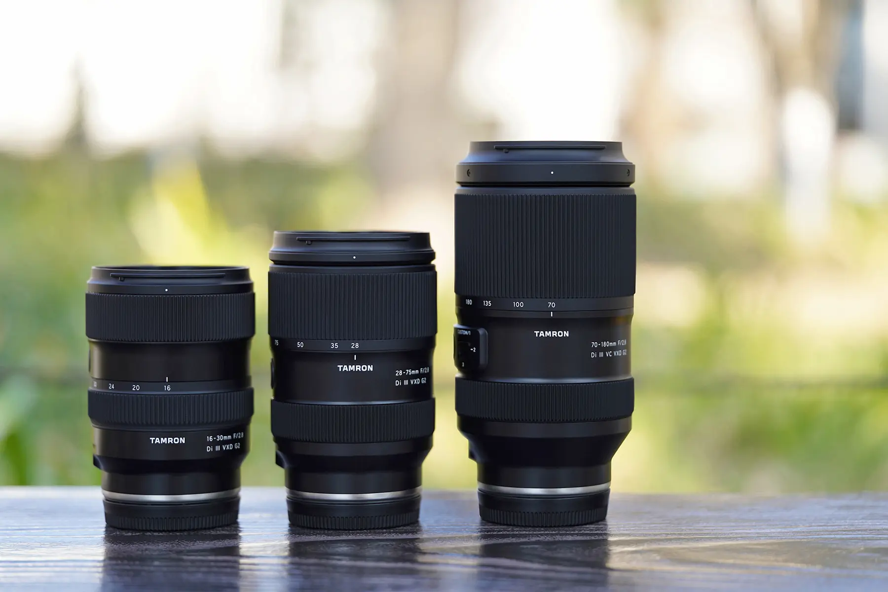 Three Tamron Di III VXD G2 lenses&mdash;a 16-30mm, 28-75mm, and 70-180mm&mdash;lined up side by side on a wooden surface with a softly blurred outdoor background, highlighting their compact design and focal range coverage.