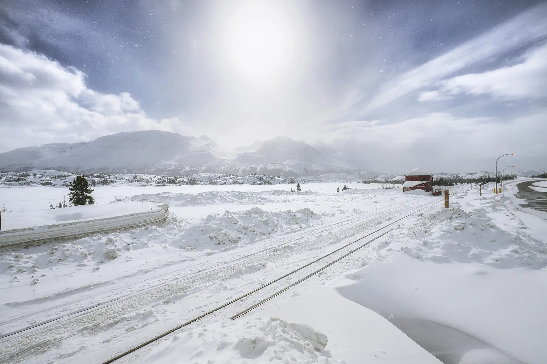 A bright winter sun hovers over a snowy mountain landscape and railroad tracks leading toward a small red building, with snowflakes drifting in the crisp air and dramatic clouds stretching across the sky.