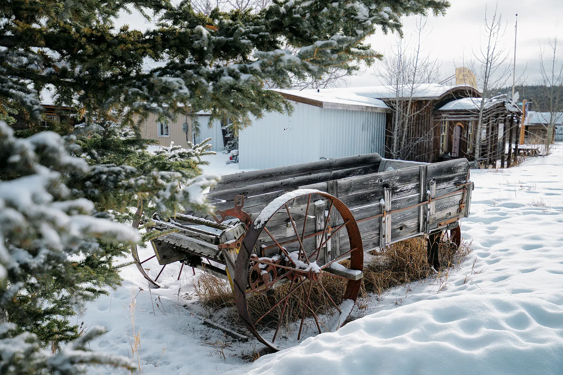 An old wooden wagon with rusted iron wheels sits partially buried in snow beside rustic buildings and evergreen trees in a quiet, snow-covered village scene.