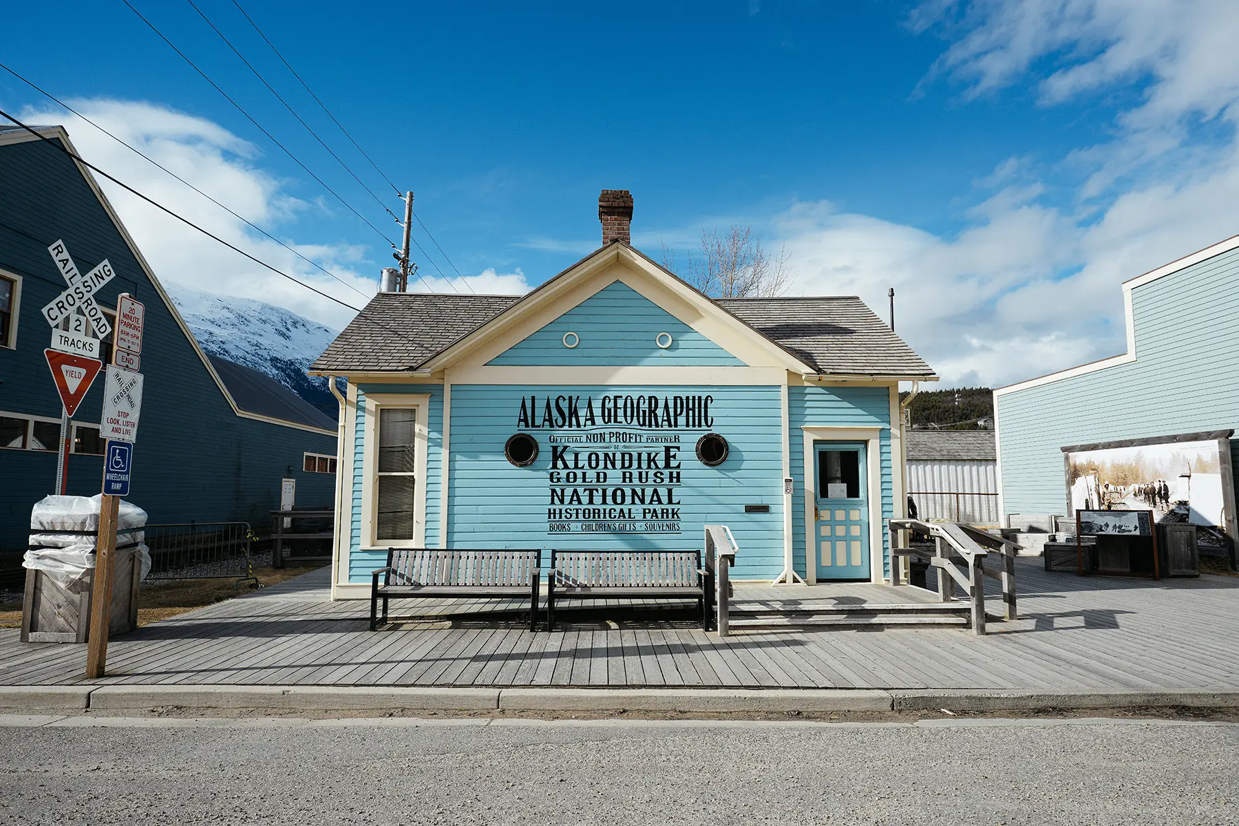 A bright blue wooden building with signage for Alaska Geographic and the Klondike Gold Rush National Historical Park stands under a clear sky, surrounded by boardwalk benches and historic rail crossing signs in a quaint Alaskan town.