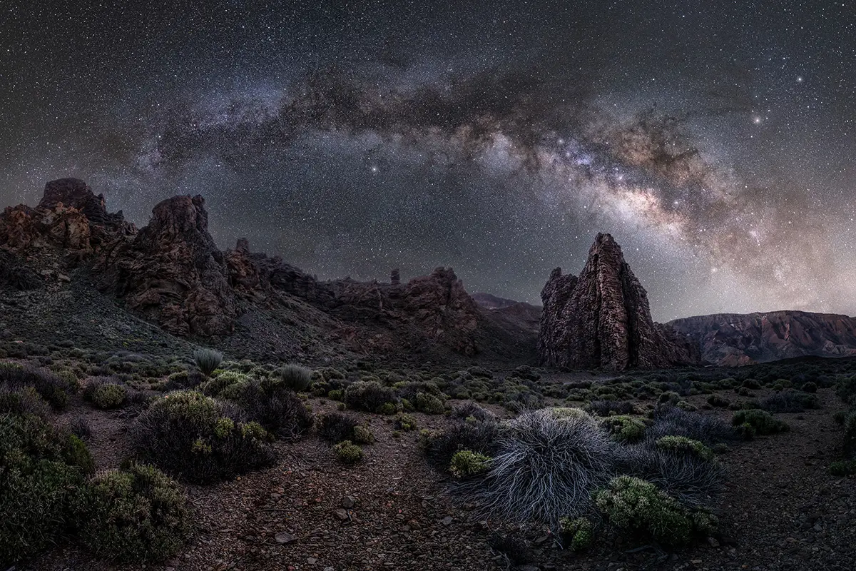 Milky Way arching over volcanic rock formations and desert vegetation under a clear, star-filled night sky