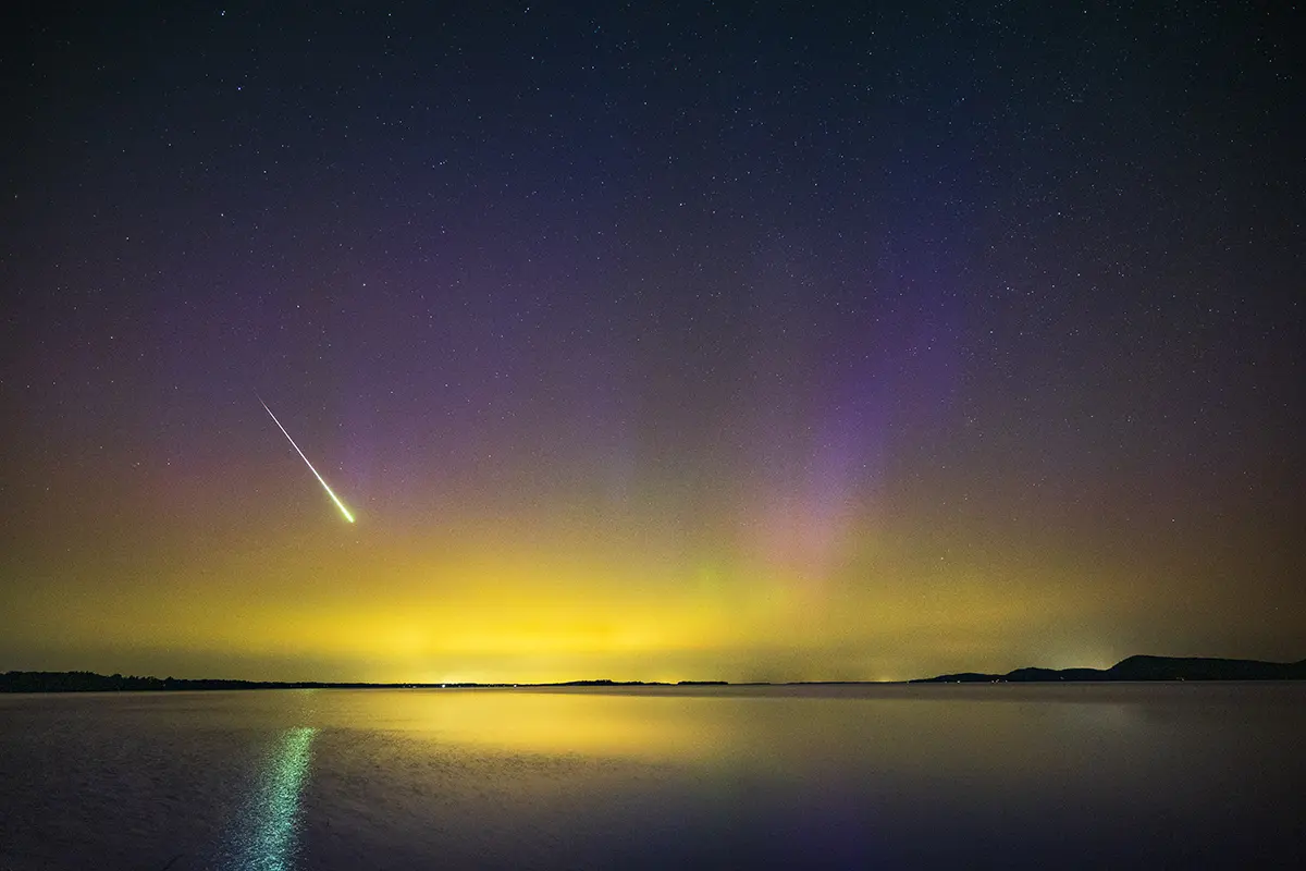 Brilliant meteor streak across a colorful aurora night sky reflected on a calm lake surface.