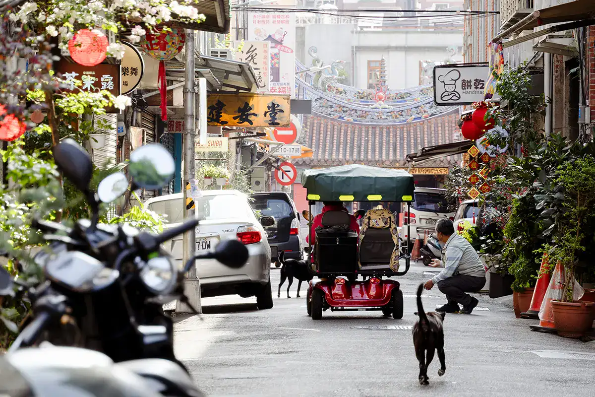 Narrow Asian street scene with scooters, market signs, and locals interacting, captured with the best lens for street photography.