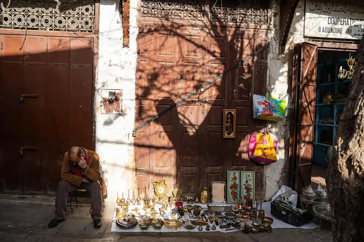 Street vendor sitting beside a display of brassware and colorful bags in front of a weathered wooden door, with tree shadows cast across the wall.