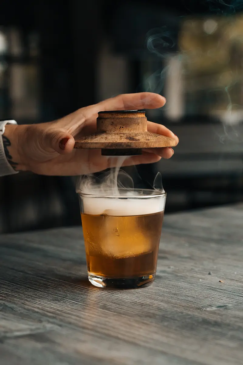 Levantamento manual de uma tampa de madeira de um copo de coquetel defumado com vapor ascendente, capturado para fotografia de alimentos e bebidas no outono.