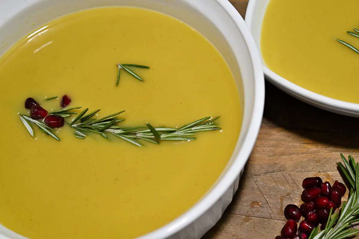 Tigela de sopa de abóbora decorada com alecrim e sementes de romã, em uma mesa rústica.