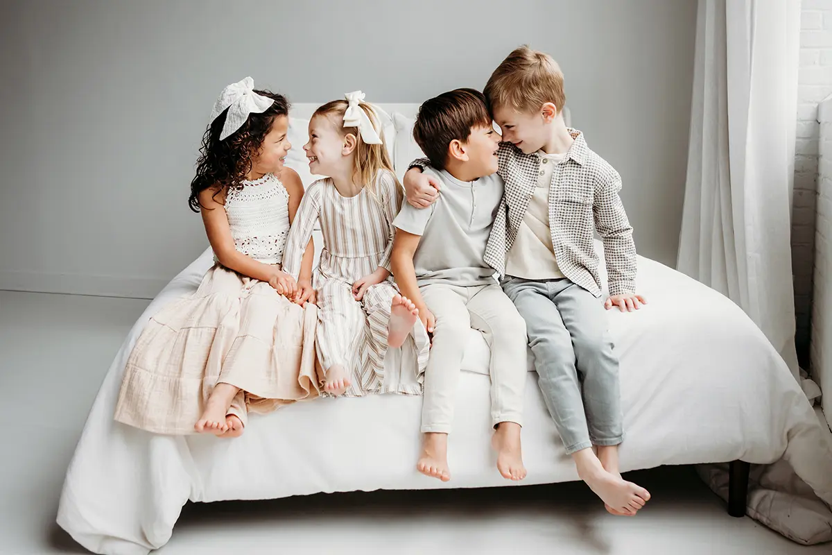 Quatre jeunes enfants assis ensemble sur un lit, souriant et interagissant au cours d'une séance photo - exemple d'objectif principal ou de zoom pour la photographie de famille.